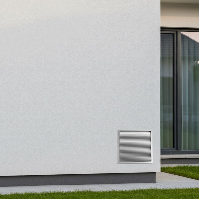 Exteriorul unei clădiri moderne de culoare albă, cu o grilă de ventilație aproape de sol și o fereastră mare parțial vizibilă în dreapta, lângă un petic de iarbă verde.